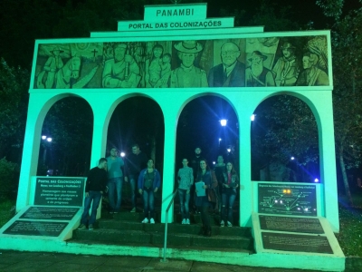 Imagem de um pórtico, com 5 pilares e uma imagem desenhada na parte superior, localizado na praça central de Panambi. A imagem foi feita à noite e, no meio do pórtico, estão posicionados em pé alguns alunos de edificações. 